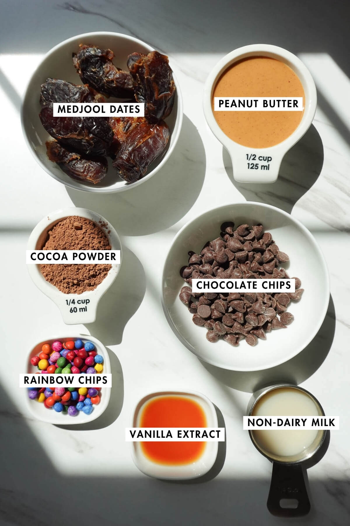 Measured ingredients to make no bake cosmic brownies including medjool dates, cocoa powder, peanut butter, chocolate chips, non-dairy milk, vanilla, and sprinkles.