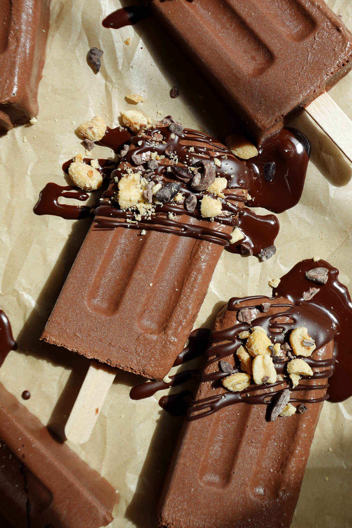 Vegan fudgesicles on a parchment-lined tray drizzled with melted chocolate and chopped peanuts on top.