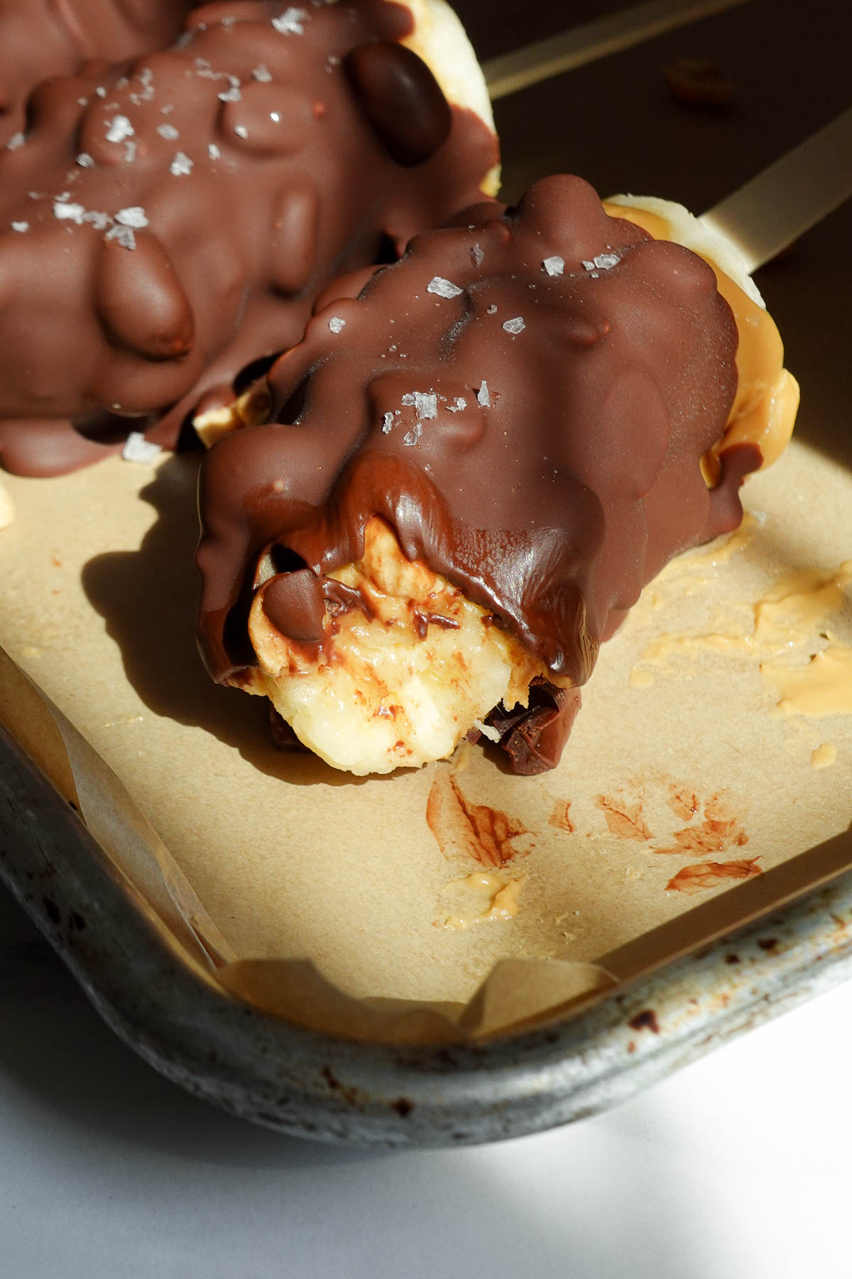 Frozen snickers banana pops with a bite showing showing layers of banana, peanut butter, peanuts, and chocolate.